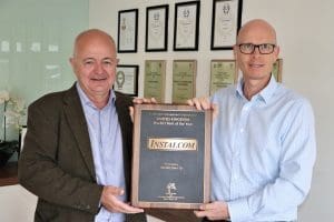 Two men standing indoors, smiling and holding a plaque that reads INSTAL.COM United Kingdom World Client of the Year. Their achievement reflects strong leadership, with framed certificates and awards displayed on the wall behind them.