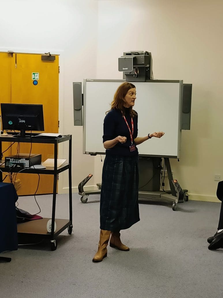 A woman stands and speaks in a classroom, gesturing with her hands during executive performance training courses. Behind her are a whiteboard and electronic equipment, with a computer workstation and wooden double doors to the left.
