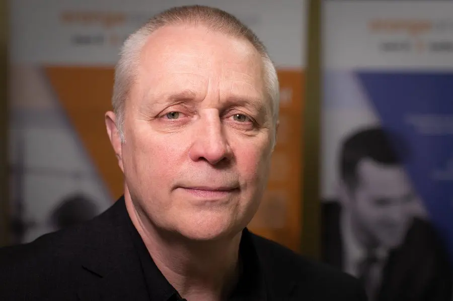 A middle-aged man with short, fair hair and blue eyes, dressed in a black shirt and blazer, looks directly at the camera with a neutral expression. The blurred background, featuring orange and blue banners, hints at an executive performance setting.