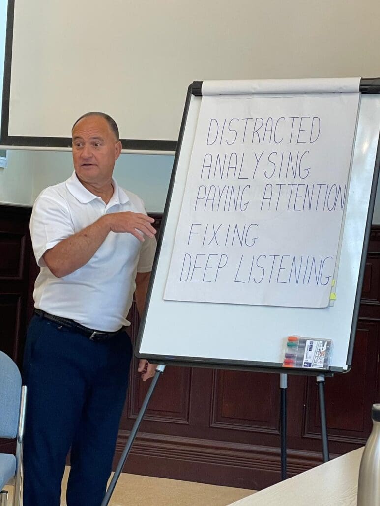 A man in a white polo shirt stands beside a flip chart with the words Distracted, Analysing, Paying Attention, Fixing, Deep Listening, gesturing as he speaks to an audience during a leadership training course.