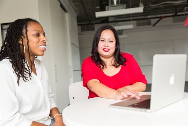 Two women sit at a round table in an office, smiling at a laptop. One wears a white blouse, the other a red top—they appear to be having a friendly discussion or collaborating on management and executive performance strategies.
