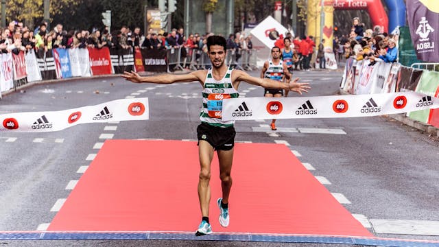 A male runner crosses the finish line of a road race with his arms outstretched, breaking the tape. Spectators line the street, whilst another runner approaches behind him—reflecting leadership and executive performance in an organised event.