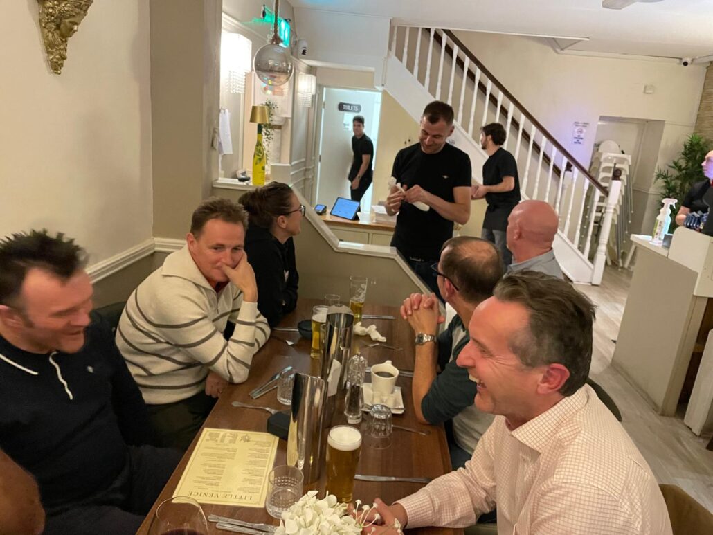 A group of people sit round a wooden table in a restaurant, smiling and talking, with drinks and menus in front of them. Demonstrating leadership, a waiter stands nearby, interacting with the group. A staircase is visible in the background.