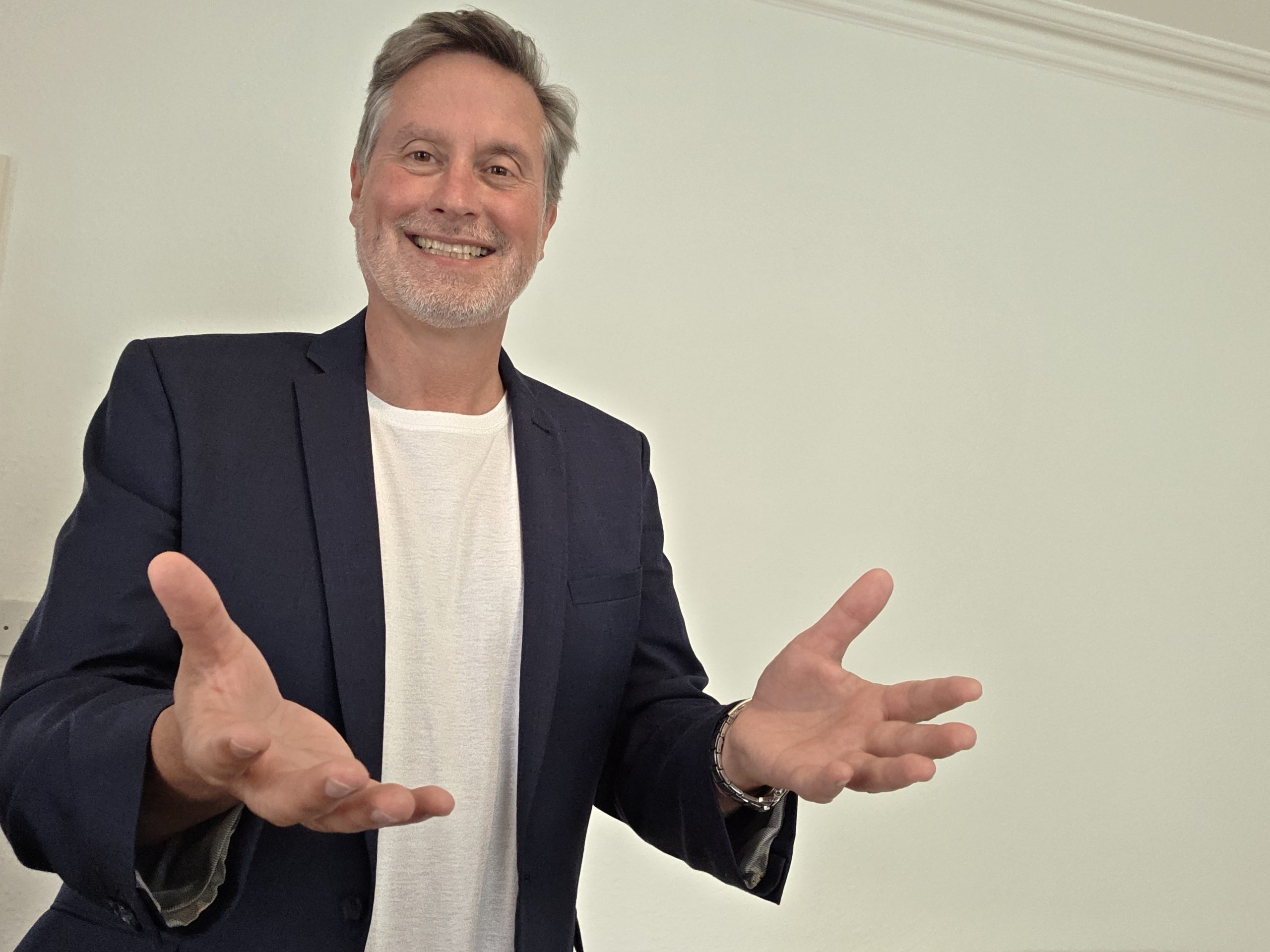 Gary Dawes smiling with grey hair and a beard wears a navy blazer over a white shirt, standing indoors with his hands open and gesturing in a friendly, welcoming manner.
